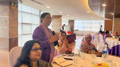 Bangladeshi woman standing up from a table holding a microphone and asking questions.