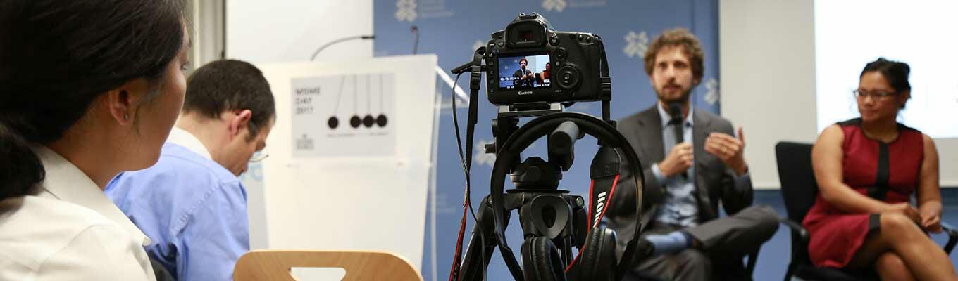 Wide image with camera on a tripod in front centre, with a man and a woman seated, blurred in the background