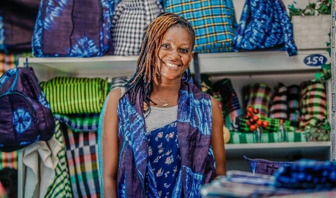 Gambian woman smiling at the camera in front of a wall of colourful fabric bags behind her on shelves